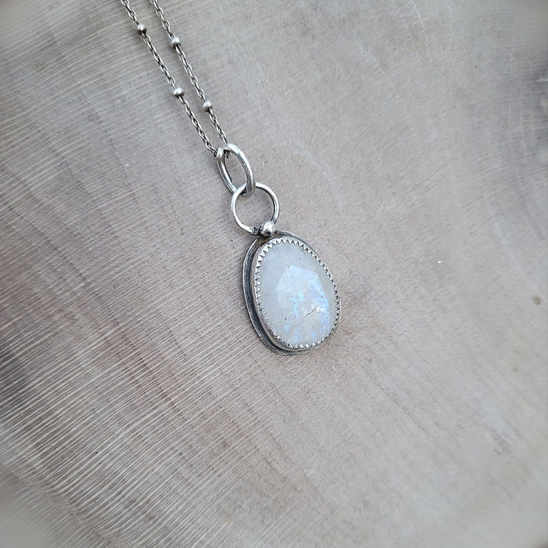 Puede incluir: Un collar de cadena de plata con un colgante de piedra lunar blanca. El colgante est&aacute; engastado en un bisel de plata y tiene un borde delicado y festoneado.
