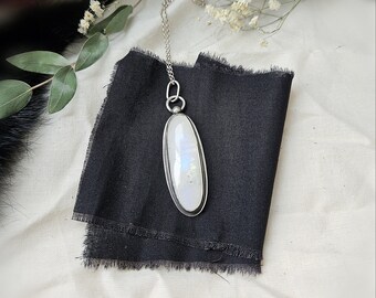 May include: A silver necklace with a large, oval, white pendant. The pendant has iridescent, rainbow-like flashes. The necklace is displayed on a black fabric with white flowers and greenery in the background.