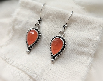 May include: A pair of silver dangle earrings with teardrop-shaped orange gemstones. The earrings have a beaded border around the gemstones.
