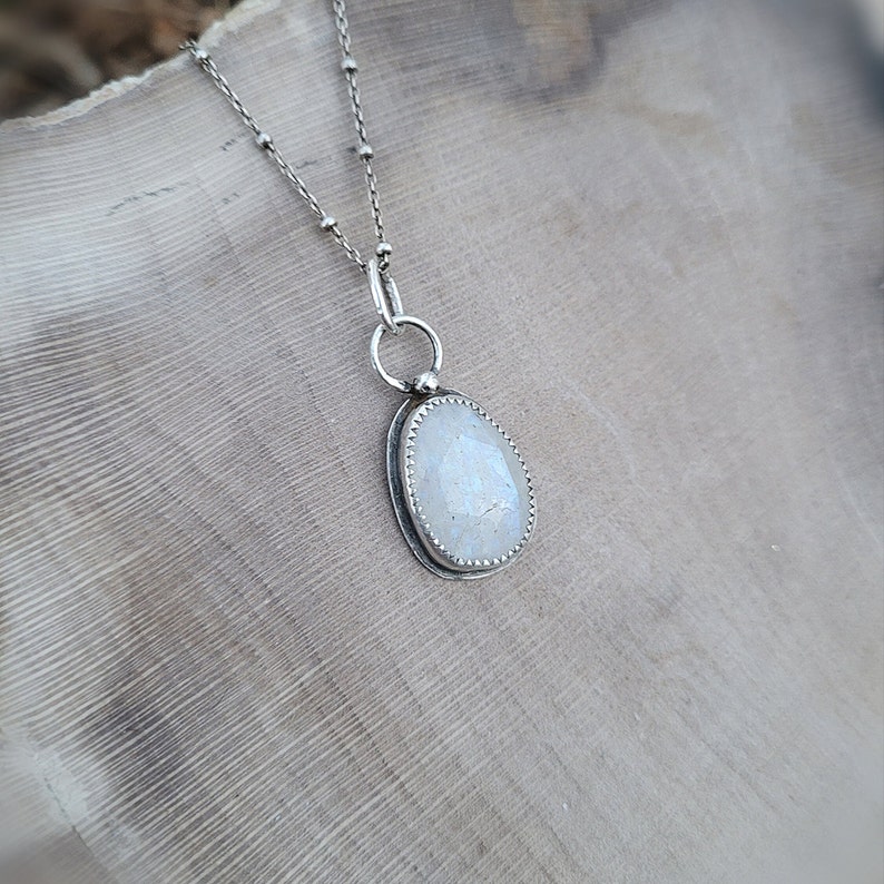 Puede incluir: Un collar de cadena de plata con un colgante de piedra de luna ovalada blanca. El colgante est&aacute; engastado en un bisel de plata con un borde festoneado.