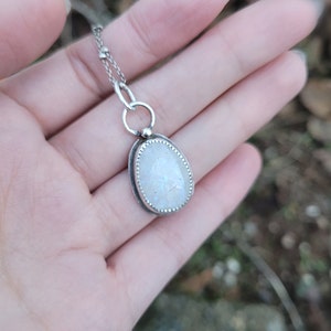 Puede incluir: Un collar de cadena de plata con un colgante de plata con una piedra de luna blanca.