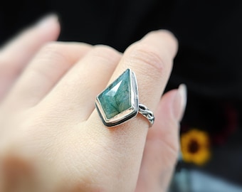 May include: A silver ring featuring a large, emerald-green gemstone cut into a kite shape. The gemstone is set in a silver bezel with a black border. The ring's band has a braided design.