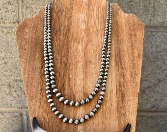 Op de afbeelding: Een zilveren kralenketting met twee strengen, tentoongesteld op een houten standaard. Onder de ketting bevindt zich een witte stierschedel met zwartgepunte hoorns. De standaard staat tegen een rustieke bakstenen muur.