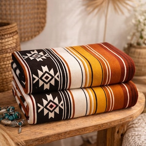 May include: Two folded blankets with a Southwestern design. The blankets feature a repeating geometric pattern in black and white, with stripes of white, yellow, orange, and brown. The blankets are stacked on a wooden table.