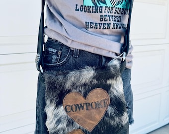 May include: A black and white cowhide crossbody bag with a brown leather heart-shaped patch that says "COWPOKE" in black lettering.