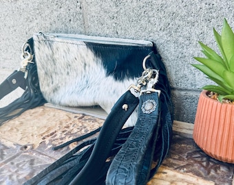 May include: A black and white cowhide clutch bag with a black fringe and a black leather strap. The strap has a silver buckle and a silver charm.