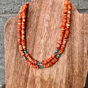 Könnte beinhalten: Eine mehrreihige Halskette mit orangefarbenen und weißen Perlen. Die Halskette hat türkisfarbene und schwarze Akzentperlen. Die Halskette wird auf einem Holzständer vor grauem Hintergrund präsentiert.