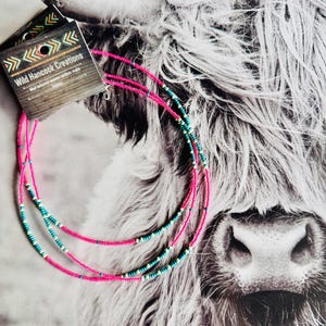 May include: Three layered beaded necklaces with pink, white, and teal beads. The necklaces are displayed on a fuzzy white and gray background.