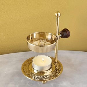May include: A gold metal incense burner with a small candle holder and a removable bowl for incense. The bowl is filled with small white incense pellets. The burner has an ornate design.
