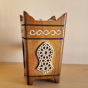 May include: A handcrafted wooden incense burner with a rich brown finish. The square-shaped burner features intricate cut-out star patterns and decorative gold-colored accents. The design includes a decorative top and small feet.
