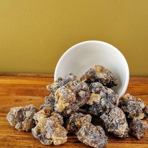 May include: A collection of irregularly shaped, amber-colored resin pieces spilling from a white bowl. The resin has a textured surface and varying shades of brown and yellow. The background is a solid mustard yellow.