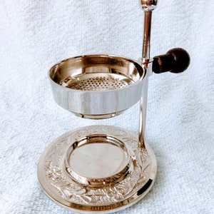 May include: Silver incense burner with a round base and a decorative design. The burner has a removable bowl with a mesh screen and a handle.