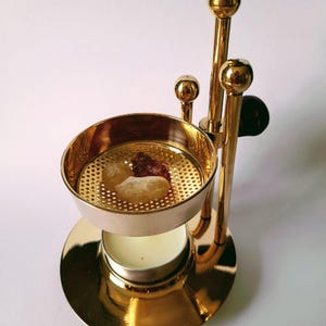 Puede incluir: Un quemador de incienso de metal dorado con una parte superior redonda y tres patas. El quemador tiene un pequeño portavelas redondo y blanco debajo de la parte superior. Hay dos piezas de incienso en la parte superior del quemador.