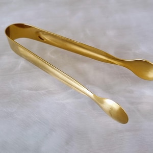May include: Gold-toned metal sugar tongs with rounded ends. The tongs are designed for grabbing and holding sugar cubes.