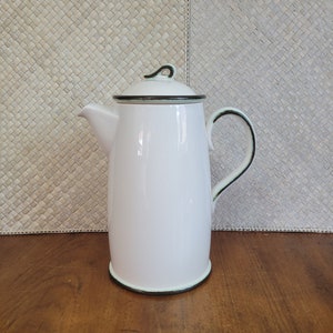 May include: White enamel coffee pot with a black rim and handle. The pot has a curved spout and a lid with a black handle. The pot is on a wooden surface with a woven background.