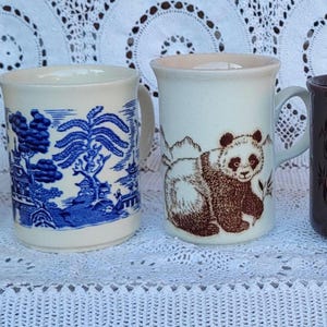 May include: Four ceramic mugs arranged in a row. The first is yellow with black rabbit illustrations and a blue rim. The second is white with a blue willow pattern. The third is white with a brown panda illustration. The fourth is dark brown with orange floral designs.