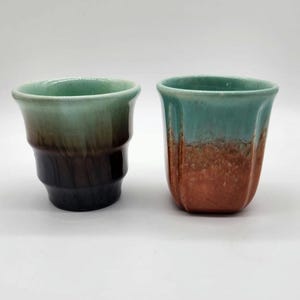 May include: Two ceramic cups with a gradient colour scheme. One cup transitions from green to brown to black. The other cup has a teal top and a brown base. Both cups have a glossy finish.