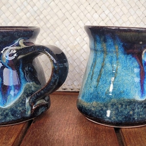 May include: Two ceramic mugs with a glossy finish featuring a blend of deep blue, turquoise, and dark purple glaze. Each mug has a wide base, a flared rim, and a curved handle on the side. The glaze shows variations in colour and texture.