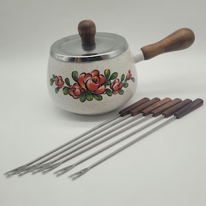 May include: A white fondue pot with a floral design and a wooden handle and lid knob. Six fondue forks with wooden handles are arranged in front of the pot. The pot is approximately 15 cm in diameter.