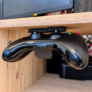 Könnte beinhalten: Ein schwarzer Videospiel-Controller, der mit einer schwarzen Kunststoffhalterung unter einem Holzregal befestigt ist. Der Controller hat eine glänzende Oberfläche, einen zentralen Anschluss und sichtbare farbige Tasten an der Oberkante.