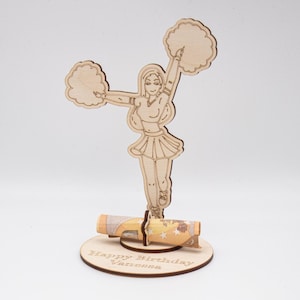 Könnte beinhalten: Eine Cheerleader-Figur aus Holz, die Pompons hält, mit einem eingerollten Geldschein in der Basis. Die Basis ist mit "Happy Birthday Vanessa" graviert.