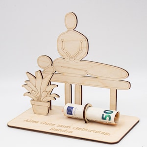 Könnte beinhalten: Ein hölzernes Geburtstagsgeschenk mit einer Person, die auf einer Massageliege liegt, und einer anderen Person, die darüber sitzt. Eine aufgerollte Banknote wird von einem Holzring gehalten. Auf der Basis steht der Text "Alles Gute zum Geburtstag, Sandra".