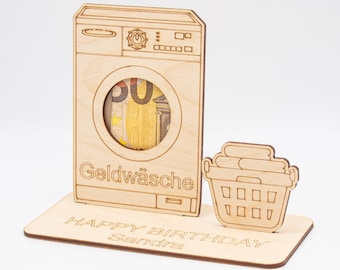 May include: A wooden money box shaped like a washing machine with a slot for money. The box has the text "Geldwäsche" on the front. The box is on a wooden base with the text "HAPPY BIRTHDAY Sandra".