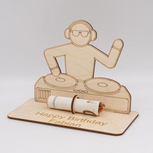Könnte beinhalten: Ein DJ-Ständer aus Holz mit einer Ausschnittfigur eines DJs, der Kopfhörer trägt und ein Mikrofon hält. Der Ständer hat zwei Plattenspieler und einen Schlitz für einen eingerollten Geldschein. Die Basis des Ständers hat den Text "Happy Birthday Fabian" eingraviert.
