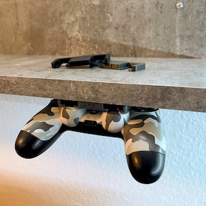 Könnte beinhalten: Ein Camouflage-Videospielcontroller ist mit einer 3D-gedruckten Halterung an einem Regal befestigt. Der Controller hängt kopfüber mit den Knöpfen nach oben.