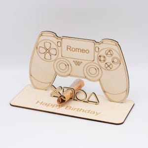 Könnte beinhalten: Ein Holzschnitt eines Videospiel-Controllers mit dem Namen "Romeo" darauf. Der Controller hält einen zusammengerollten Geldschein. Die Basis des Controllers trägt die Aufschrift "Happy Birthday".