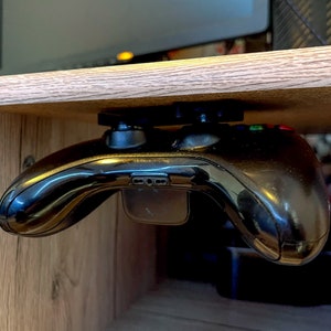 Könnte beinhalten: Ein schwarzer Videospielcontroller ist an der Unterseite eines Holzregals befestigt. Der Controller hängt kopfüber mit den Knöpfen nach oben.