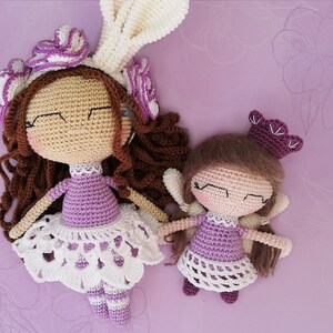 May include: Two crocheted dolls wearing purple dresses with white lace trim. The larger doll has brown hair and a purple flower crown. The smaller doll has a purple crown. The dolls are on a purple background with the words "Violet" and "Amethyst" written on either side.