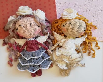 May include: Two crocheted angel dolls with white wings and ruffled skirts. One doll has brown hair with a white flower crown and a gray skirt. The other doll has blonde hair with a white flower crown and a white skirt.