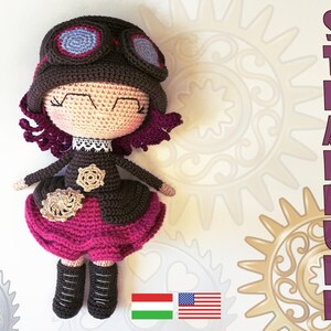 Puede incluir: Una muñeca de crochet con cabello castaño, un sombrero marrón con gafas, un vestido marrón y rosa y botas marrones. La muñeca está de pie sobre un fondo blanco con un patrón de engranajes y corazones. El texto "STEAMPUNK" está escrito verticalmente en letras rosas en el lado derecho de la imagen.