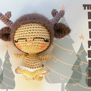 May include: A crocheted reindeer doll with brown antlers, a brown hat, and a yellow skirt. The doll has a smiling face with closed eyes and rosy cheeks. The doll is sitting in front of a background of green Christmas trees.