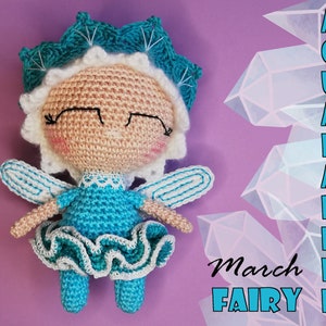 May include: A crocheted fairy doll with blue and white wings, a white dress with a blue ruffle, and a blue and white crown. The doll has a pink face with black eyes and a closed mouth. The background is purple with a crystal design and the text "Aquamarine March Fairy".