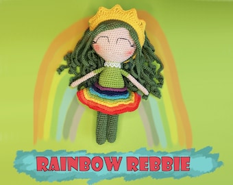 May include: A crocheted doll with green hair and a rainbow skirt, wearing a yellow crown. The doll is posed against a rainbow background with the text "RAINBOW REBBIE".