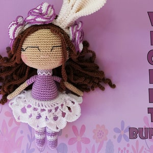 May include: Crocheted doll with brown hair, a purple dress, and a white lace skirt. The doll is wearing a white bunny ear headband with purple flowers. The doll is lying on a purple background with the text "VIOLET BUNNY" in pink.