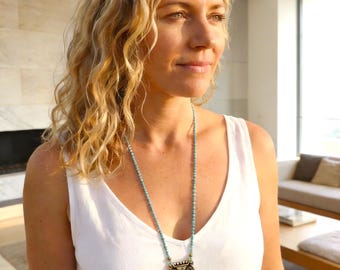 May include: A long necklace featuring small turquoise-coloured beads, a rectangular metallic pendant with intricate etched patterns, a square blue stone, and a teardrop-shaped blue stone pendant hanging from a white sleeveless top.