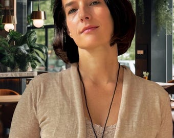 May include: A woman in a beige cardigan and camisole, with a black pendant necklace. The background shows a restaurant interior with hanging plants and pendant lights, creating a natural ambiance.