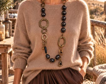 May include: A beige long-sleeved knit jumper paired with a dark brown skirt and a long necklace featuring dark spherical beads with white accents, interspersed with large, textured, circular brass-toned metal links and a small dangling charm.
