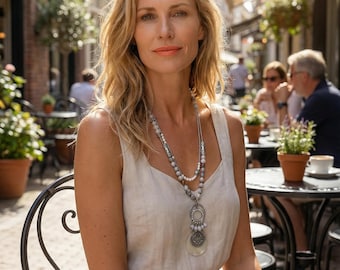 May include: A layered necklace featuring small grey and white beads, silver-toned metal accents, and a circular pendant with a textured centre and a smooth, round white disc base, worn with a sleeveless beige dress with a scoop neckline.