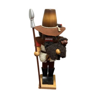May include: A wooden nutcracker figure wearing a brown hat and holding a silver-tipped spear. The figure has a white faux fur vest, dark brown trousers, and black boots. It holds a small, dark brown, fuzzy sheep figure in its arms.