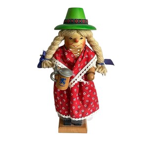 May include: A wooden nutcracker figurine wearing a green hat with a floral band, blonde yarn braids, and a red dress with a white floral pattern and lace trim. The figure holds a small wooden beer stein featuring a blue lion emblem on the front.
