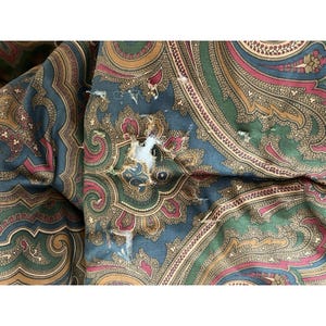 Op de afbeelding: Een textielstof met een meerkleurig paisley-patroon in tinten donkergroen, dieprood en goud. Het materiaal vertoont zichtbare gebruikssporen, waaronder gerafelde randen en een scheur in het midden die de witte binnenvoering blootlegt.
