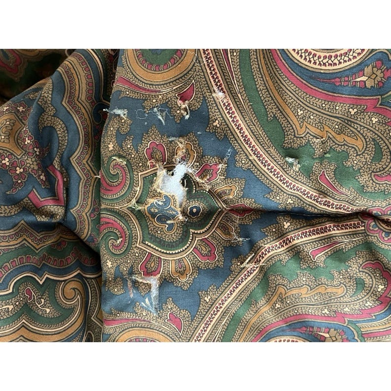 Op de afbeelding: Een textielstof met een meerkleurig paisley-patroon in tinten donkergroen, dieprood en goud. Het materiaal vertoont zichtbare gebruikssporen, waaronder gerafelde randen en een scheur in het midden die de witte binnenvoering blootlegt.