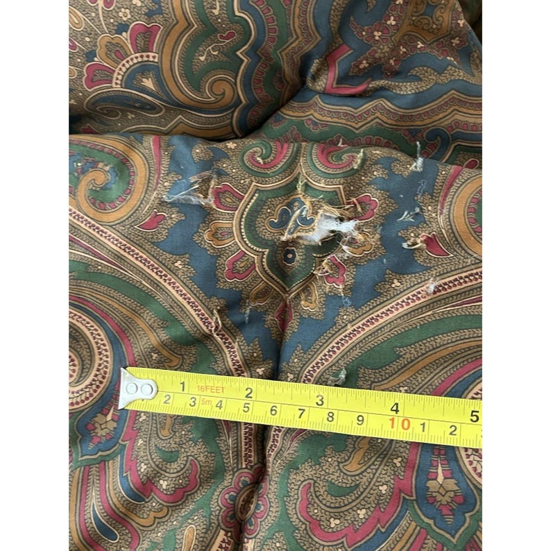 Op de afbeelding: Een stoffen kussen met een veelkleurig paisley-patroon in tinten blauwgroen, groen, goud en bordeauxrood. Een geel meetlint ligt over het oppervlak en toont een scheur in de stof van ongeveer 3,8 cm lang.
