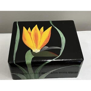 May include: A black wooden box with a hand-painted yellow and orange flower design. Green leaves and stems surround the flower. The box has a glossy finish and is likely used for storage or display.