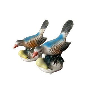 May include: Two ceramic bird figurines with blue and brown painted details, perched on a base with a yellow pear. The birds have red beaks and are facing different directions.