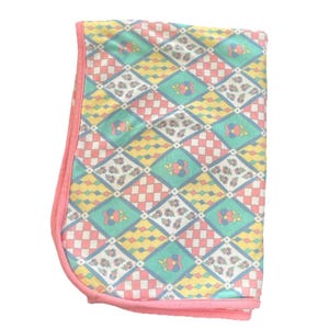 May include: A folded baby blanket with a colorful geometric pattern. The blanket features a repeating design of diamonds, squares, and floral motifs in shades of pink, yellow, green, and blue. The edges are trimmed with a solid pink border.